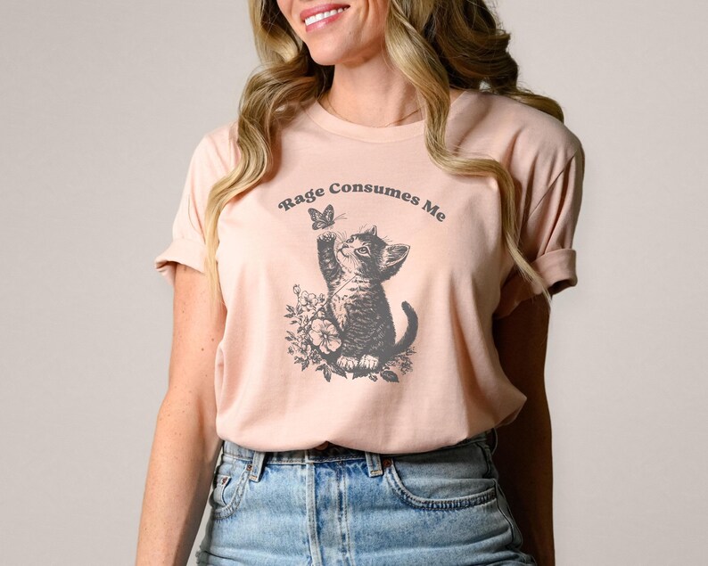 May include: A light pink t-shirt featuring a graphic of a kitten reaching for a butterfly, surrounded by floral elements. The text "Rage Consumes Me" is arched above the kitten. The shirt is made of soft material.