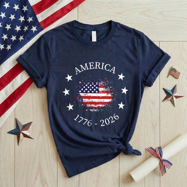 Może przedstawiać: Granatowa koszulka z nadrukiem w postaci postarzonej flagi amerykańskiej. Koszulka ma napis "AMERICA" nad flagą i lata "1776 - 2026" pod nią, otoczone białymi gwiazdami. W tle widoczna jest flaga amerykańska.