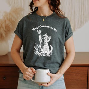 May include: Dark gray t-shirt featuring a white graphic of a kitten reaching for a butterfly, surrounded by flowers. The text "Rage Consumes Me" is arched above the graphic. The shirt has rolled-up sleeves.