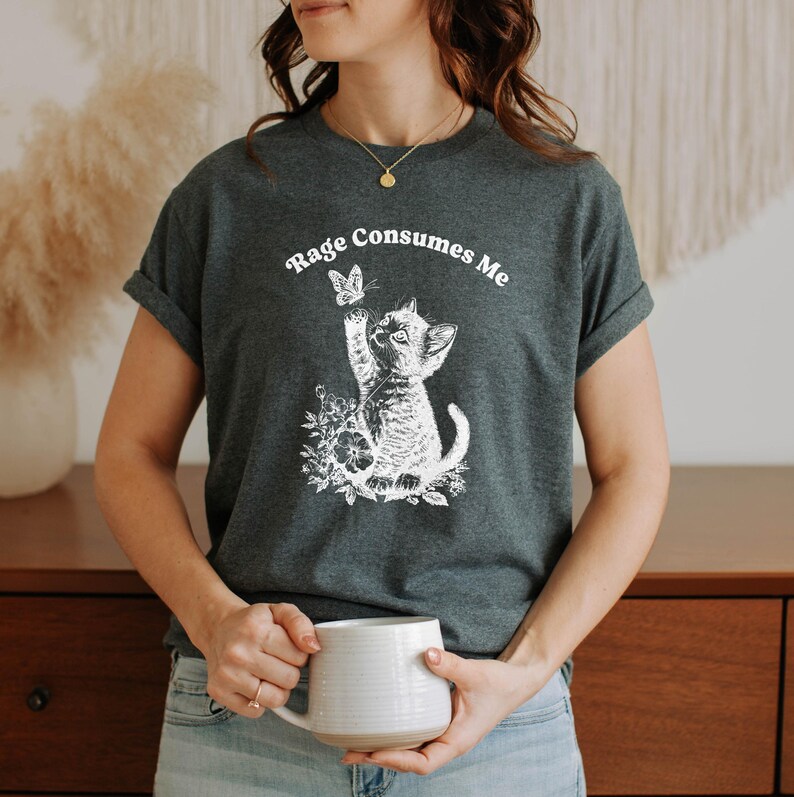 May include: Dark gray t-shirt featuring a white graphic of a kitten reaching for a butterfly, surrounded by flowers. The text "Rage Consumes Me" is arched above the graphic. The shirt has rolled-up sleeves.