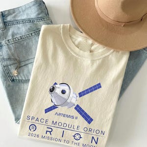 May include: Cream-colored t-shirt featuring a graphic of the Artemis II Orion space module with blue solar panels. The text on the shirt reads "SPACE MODULE ORION 2026 MISSION TO THE MOON." A light brown hat and light blue jeans are also in the image.