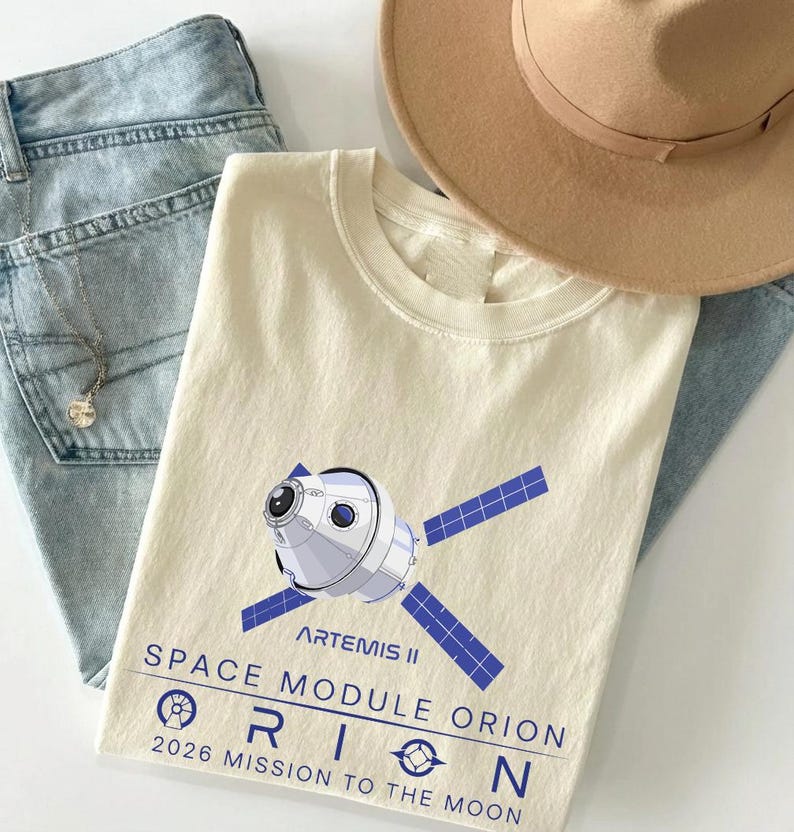 May include: Cream-colored t-shirt featuring a graphic of the Artemis II Orion space module with blue solar panels. The text on the shirt reads "SPACE MODULE ORION 2026 MISSION TO THE MOON." A light brown hat and light blue jeans are also in the image.