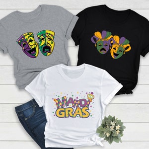 May include: Three t-shirts with Mardi Gras designs. One grey t-shirt features two purple, green, and gold masks. One black t-shirt features two purple, green, and gold jester masks. One white t-shirt features the words "Mardi Gras" in purple, green, and gold.