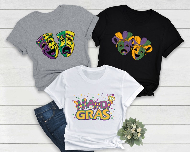 May include: Three t-shirts with Mardi Gras designs. One grey t-shirt features two purple, green, and gold masks. One black t-shirt features two purple, green, and gold jester masks. One white t-shirt features the words "Mardi Gras" in purple, green, and gold.