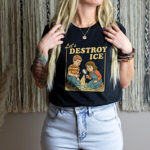 May include: Black t-shirt with the text "Let's Destroy Ice" and a graphic of two children experimenting with ice. The children are wearing vintage-style clothing. The shirt is paired with light blue denim jeans.