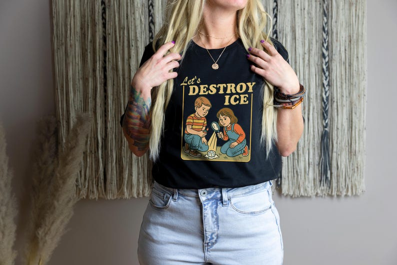 May include: Black t-shirt with the text "Let's Destroy Ice" and a graphic of two children experimenting with ice. The children are wearing vintage-style clothing. The shirt is paired with light blue denim jeans.