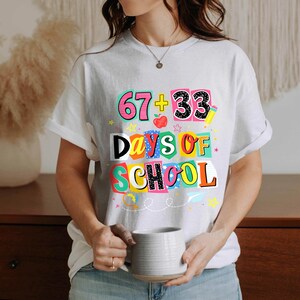 May include: White t-shirt with a colorful graphic that reads "67 + 33 Days of School." The design includes numbers, an apple, and stars. The shirt is short-sleeved and has a casual fit.
