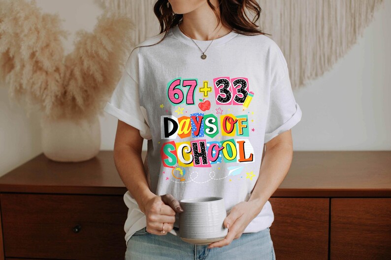 May include: White t-shirt with a colorful graphic that reads "67 + 33 Days of School." The design includes numbers, an apple, and stars. The shirt is short-sleeved and has a casual fit.