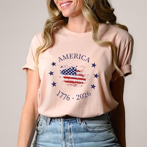 Może przedstawiać: Jasnor&oacute;żowa koszulka z postarzanym wzorem amerykańskiej flagi, napisem "AMERICA" i latami "1776 - 2026". Wz&oacute;r otoczony jest niebieskimi gwiazdami. Koszulka jest noszona przez osobę.