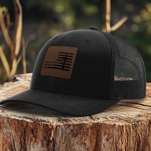 May include: Black trucker hat with a mesh back and a curved brim. The front features a brown leather patch with a flag design and the words "MAN OF GOD, HUSBAND, DAD". The hat is sitting on a wooden stump.