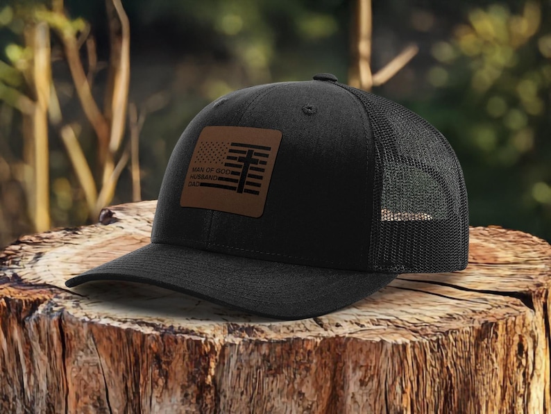 May include: Black trucker hat with a mesh back and a curved brim. The front features a brown leather patch with a flag design and the words "MAN OF GOD, HUSBAND, DAD". The hat is sitting on a wooden stump.