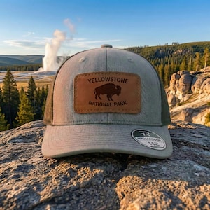 May include: A gray and olive green trucker hat with a leather patch featuring a bison and the text "YELLOWSTONE NATIONAL PARK". The hat has a mesh back and a curved brim. The Richardson 112 logo is visible.