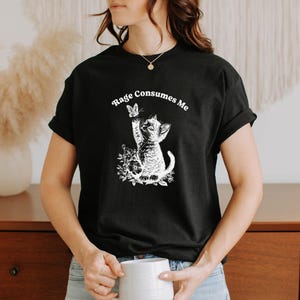 May include: Black t-shirt with a white graphic of a kitten reaching for a butterfly, surrounded by floral elements. The text "Rage Consumes Me" is arched above the graphic. The shirt is short-sleeved.