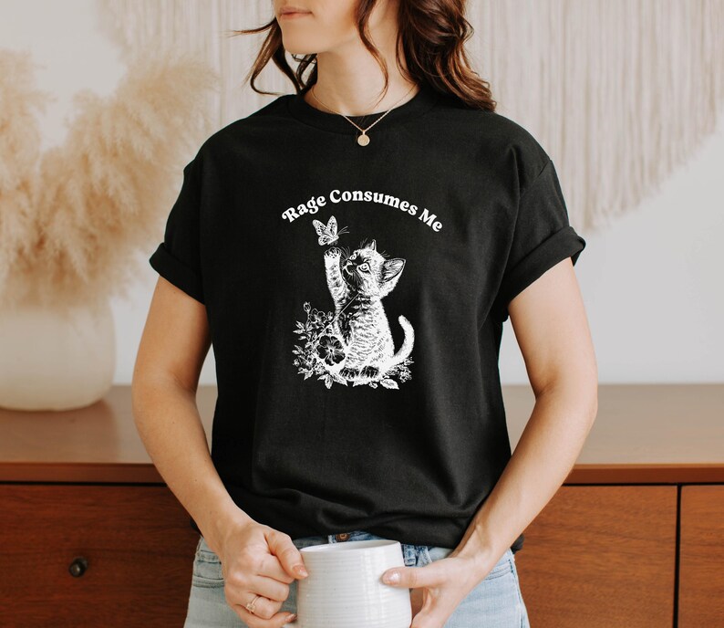 May include: Black t-shirt with a white graphic of a kitten reaching for a butterfly, surrounded by floral elements. The text "Rage Consumes Me" is arched above the graphic. The shirt is short-sleeved.