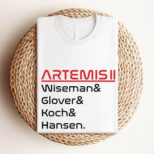 Pode incluir: Camiseta branca com o texto vermelho "ARTEMIS II" e os nomes Wiseman, Glover, Koch e Hansen em preto. A camiseta est&aacute; dobrada e colocada sobre uma superf&iacute;cie redonda de vime castanho claro.
