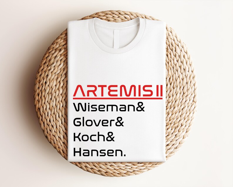 Pode incluir: Camiseta branca com o texto vermelho "ARTEMIS II" e os nomes Wiseman, Glover, Koch e Hansen em preto. A camiseta est&aacute; dobrada e colocada sobre uma superf&iacute;cie redonda de vime castanho claro.