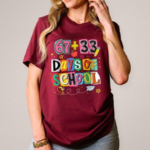 May include: Burgundy t-shirt with a colorful graphic that reads "67 + 33 Days of School." The design includes an apple, pencil, stars, and a paper airplane. The shirt is short-sleeved.