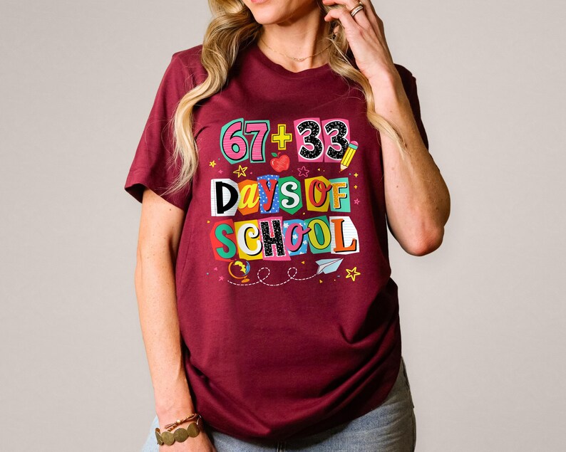 May include: Burgundy t-shirt with a colorful graphic that reads "67 + 33 Days of School." The design includes an apple, pencil, stars, and a paper airplane. The shirt is short-sleeved.