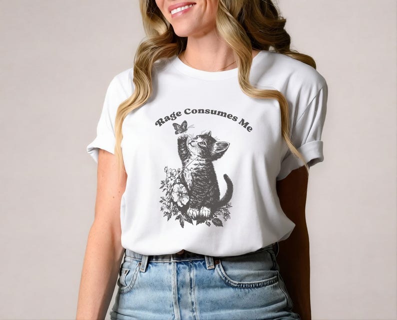 May include: White t-shirt featuring a black and white illustration of a kitten reaching for a butterfly. The text "Rage Consumes Me" is arched above the image. The shirt is worn by a person in blue jeans.