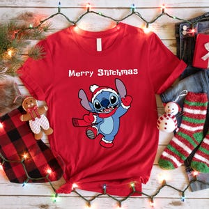 Peut inclure: Un t-shirt rouge avec le texte "Merry Stitchmas" et un personnage de dessin anim&eacute; portant un bonnet et une &eacute;charpe de P&egrave;re No&euml;l. L'image est festive, avec des d&eacute;corations de No&euml;l, un coffret cadeau et des chaussettes.