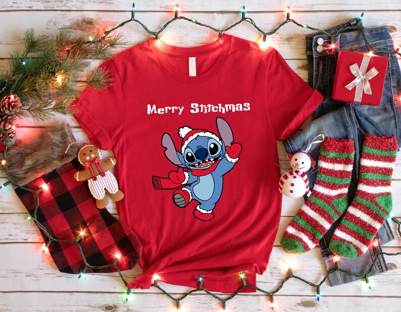 Peut inclure: Un t-shirt rouge avec le texte "Merry Stitchmas" et un personnage de dessin anim&eacute; portant un bonnet et une &eacute;charpe de P&egrave;re No&euml;l. L'image est festive, avec des d&eacute;corations de No&euml;l, un coffret cadeau et des chaussettes.