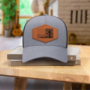 May include: A gray trucker hat with a black mesh back and a brown leather patch. The patch features an American flag design and the words "MAN OF GOD, HUSBAND, DAD" in white text. The hat is sitting on a wooden table.