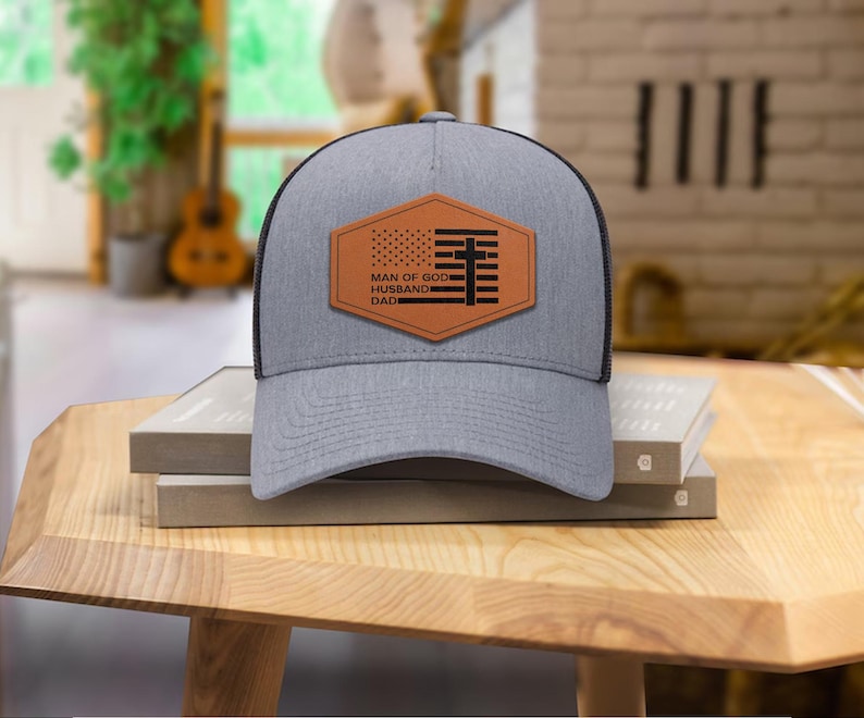 May include: A gray trucker hat with a black mesh back and a brown leather patch. The patch features an American flag design and the words "MAN OF GOD, HUSBAND, DAD" in white text. The hat is sitting on a wooden table.