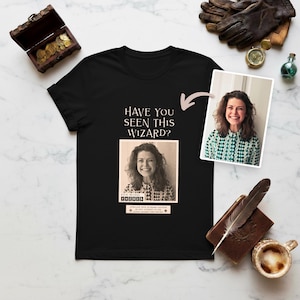 May include: Black t-shirt with the text "HAVE YOU SEEN THIS WIZARD?" and a sepia-toned image of a woman. A second image of the woman in a green and white patterned shirt is also present. Additional props include a treasure chest, compass, and a small bottle.