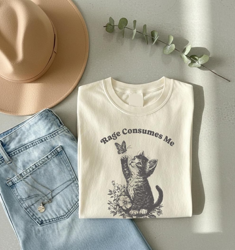 May include: Cream-colored t-shirt with the text "Rage Consumes Me" and a graphic of a kitten reaching for a butterfly. The shirt is paired with light blue jeans and a tan hat.