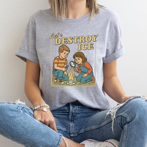 May include: A light gray t-shirt featuring a retro illustration of two children with magnifying glasses, melting a cube of ice. The text "Let's DESTROY ICE" is above the children in a yellow box.