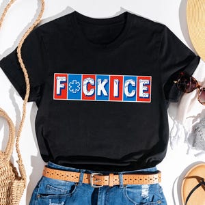 May include: Black t-shirt with the text "F*CK ICE" in a red, white, and blue design. The shirt is styled with a straw hat, sunglasses, and a woven bag.