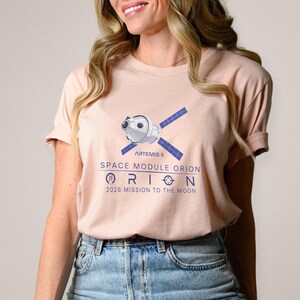 May include: A light pink t-shirt featuring a graphic of a space module with blue solar panels and the text "ARTEMIS II SPACE MODULE ORION 2026 MISSION TO THE MOON" in blue. The shirt has short sleeves.
