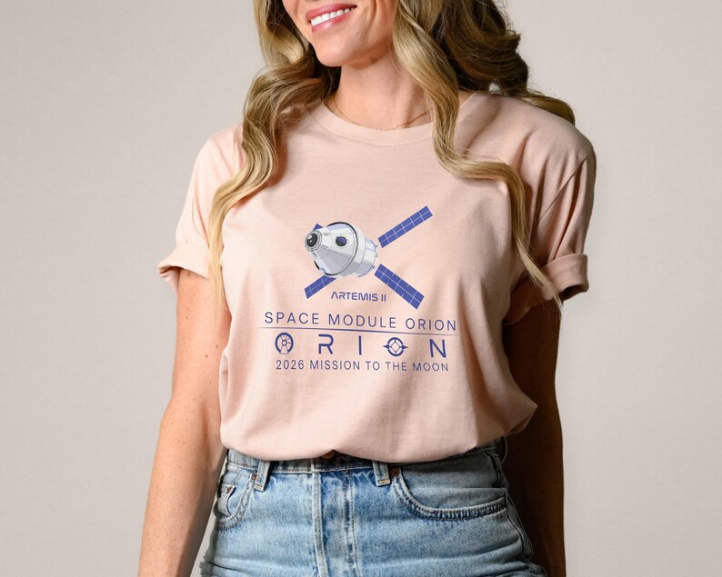 May include: A light pink t-shirt featuring a graphic of a space module with blue solar panels and the text "ARTEMIS II SPACE MODULE ORION 2026 MISSION TO THE MOON" in blue. The shirt has short sleeves.