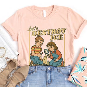 May include: A light pink t-shirt with the text "Let's DESTROY ICE" and a retro illustration of two children. One child holds a magnifying glass, and the other holds a lighter. The shirt is laid flat on a white surface.