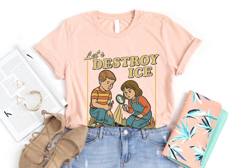 May include: A light pink t-shirt with the text "Let's DESTROY ICE" and a retro illustration of two children. One child holds a magnifying glass, and the other holds a lighter. The shirt is laid flat on a white surface.