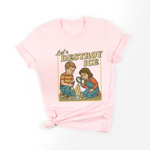 May include: Pink t-shirt featuring a retro-style illustration. The graphic shows two children kneeling, examining ice with a magnifying glass. Text above reads "Let's DESTROY ICE" in a vintage font. The children are wearing overalls and a striped shirt.
