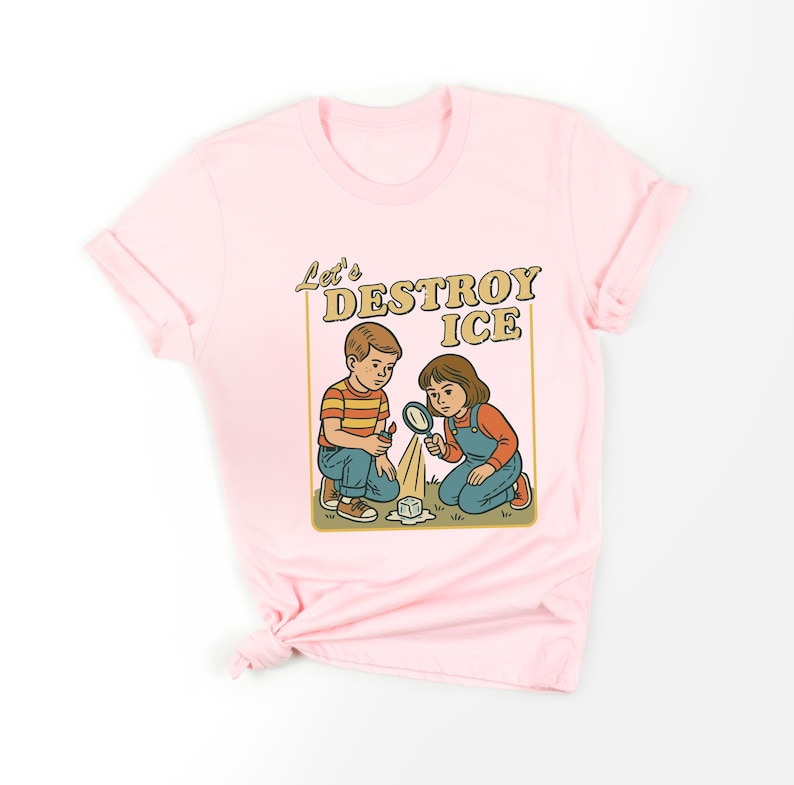 May include: Pink t-shirt featuring a retro-style illustration. The graphic shows two children kneeling, examining ice with a magnifying glass. Text above reads "Let's DESTROY ICE" in a vintage font. The children are wearing overalls and a striped shirt.