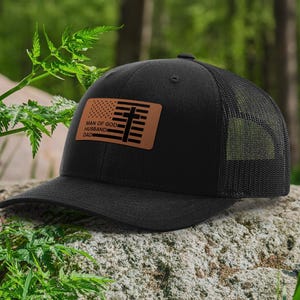 May include: Black trucker hat with a mesh back and a curved brim. The hat features a brown leather patch with an American flag design and the words "MAN OF GOD HUSBAND DAD".