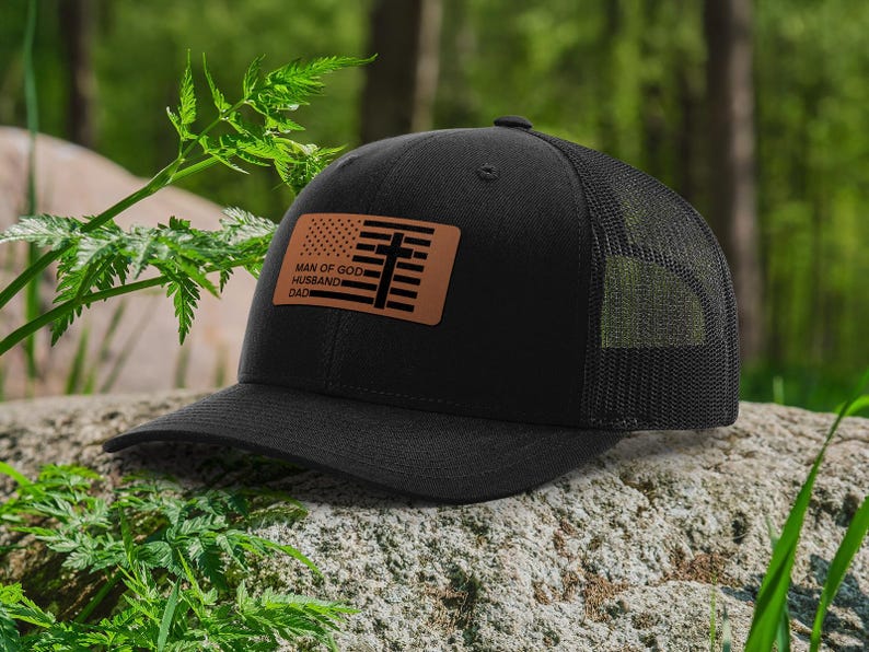 May include: Black trucker hat with a mesh back and a curved brim. The hat features a brown leather patch with an American flag design and the words "MAN OF GOD HUSBAND DAD".