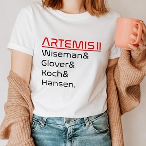 Pode incluir: Camiseta branca com o texto vermelho "ARTEMIS II" acima dos nomes Wiseman, Glover, Koch e Hansen em preto. A pessoa est&aacute; usando jeans azuis e um cardigan bege, segurando uma caneca cor de p&ecirc;ssego.