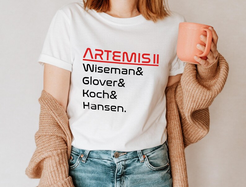 Pode incluir: Camiseta branca com o texto vermelho "ARTEMIS II" acima dos nomes Wiseman, Glover, Koch e Hansen em preto. A pessoa est&aacute; usando jeans azuis e um cardigan bege, segurando uma caneca cor de p&ecirc;ssego.