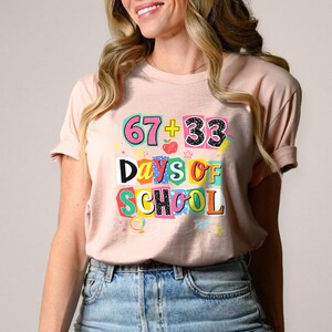 May include: A light pink t-shirt with the text "67 + 33 Days of School" in colorful block letters. The graphic includes an apple and stars. The shirt is paired with blue jeans.