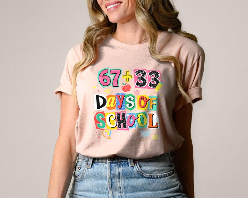 May include: A light pink t-shirt with the text "67 + 33 Days of School" in colorful block letters. The graphic includes an apple and stars. The shirt is paired with blue jeans.