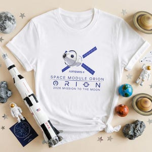May include: White t-shirt with a graphic of a space module and the text "Artemis II Space Module Orion 2026 Mission to the Moon." The image is surrounded by space-themed decorations, including a model rocket, planets, and stars.
