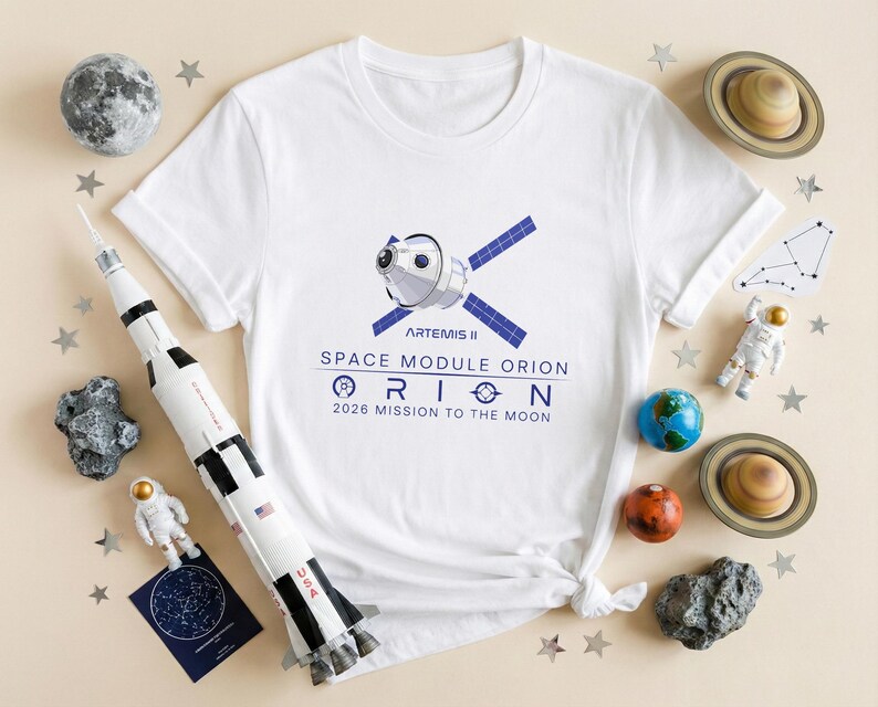 May include: White t-shirt with a graphic of a space module and the text "Artemis II Space Module Orion 2026 Mission to the Moon." The image is surrounded by space-themed decorations, including a model rocket, planets, and stars.