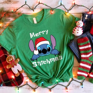 Peut inclure: T-shirt vert avec un dessin de Stitch portant un bonnet de P&egrave;re No&euml;l et l'inscription "Merry Stitchmas". L'image comprend un jean bleu, des chaussettes ray&eacute;es rouges et vertes, un coffret cadeau rouge et un ornement de bonhomme de neige.