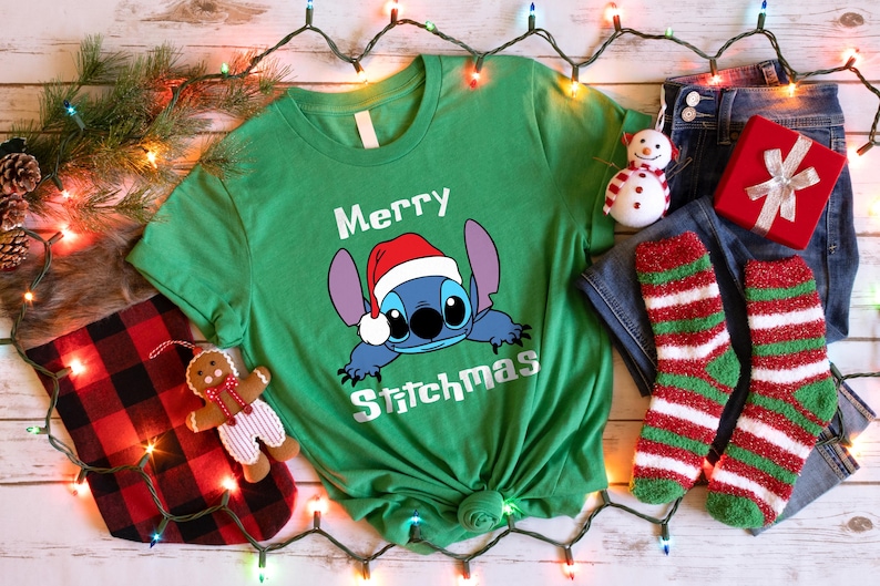Peut inclure: T-shirt vert avec un dessin de Stitch portant un bonnet de P&egrave;re No&euml;l et l'inscription "Merry Stitchmas". L'image comprend un jean bleu, des chaussettes ray&eacute;es rouges et vertes, un coffret cadeau rouge et un ornement de bonhomme de neige.