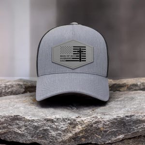 May include: A gray trucker hat with a black mesh back. The front features a patch with an American flag design and the words "MAN OF GOD, HUSBAND, DAD". The hat is sitting on a rock.