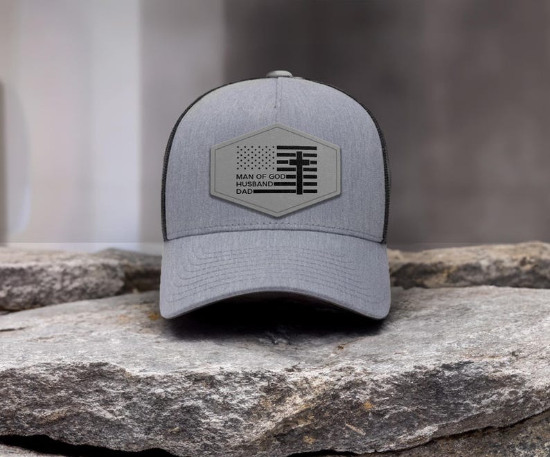 May include: A gray trucker hat with a black mesh back. The front features a patch with an American flag design and the words "MAN OF GOD, HUSBAND, DAD". The hat is sitting on a rock.