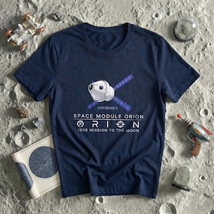 May include: Navy blue t-shirt featuring a graphic of the Artemis II Space Module Orion with text that reads "2026 Mission to the Moon." The shirt is surrounded by toy space-themed items, including a toy rocket and a lunar rover.